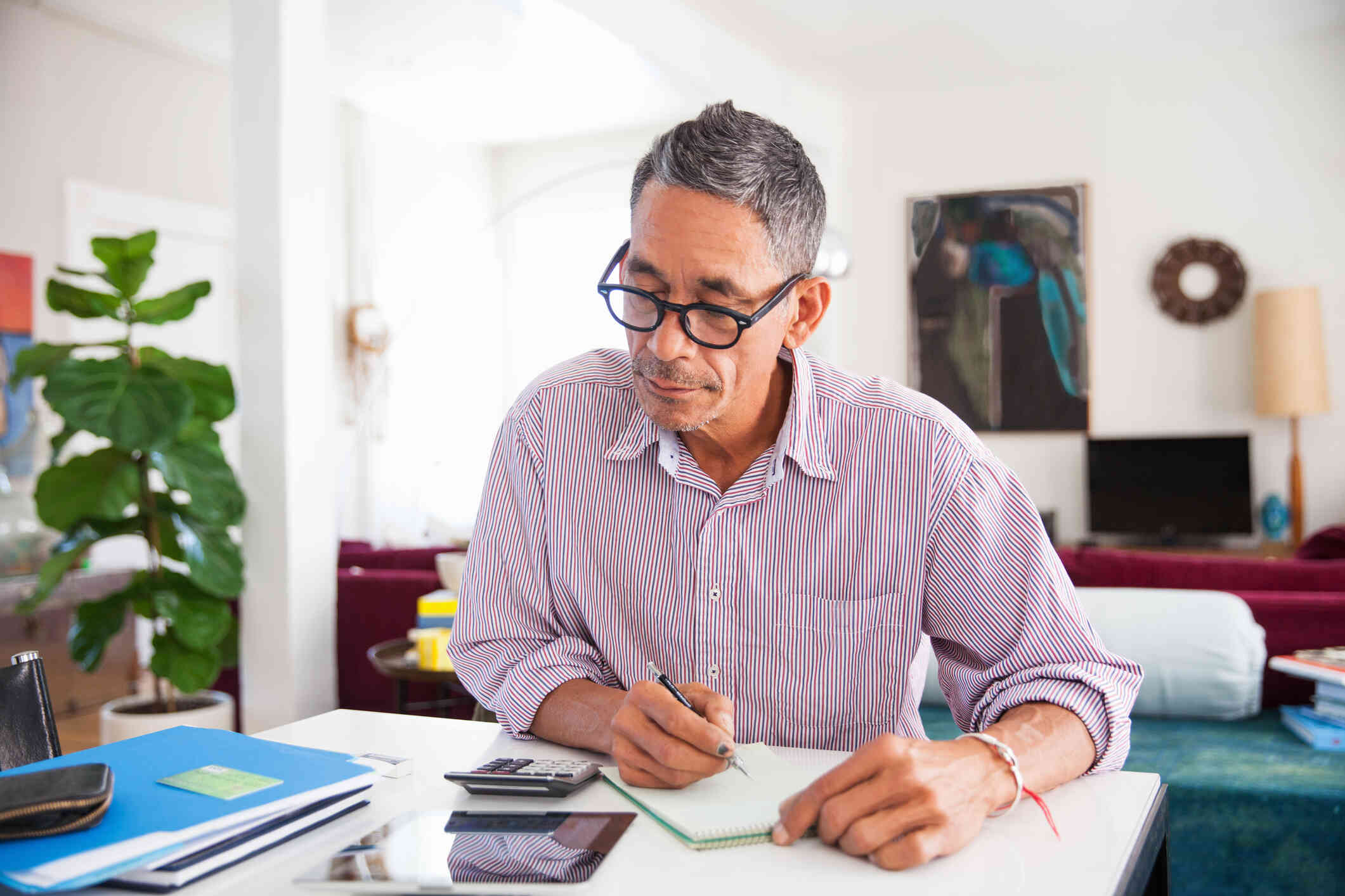 A man with glasses sits at a table in a house and writes in a notebook as a calculator and papers lay in front of him.